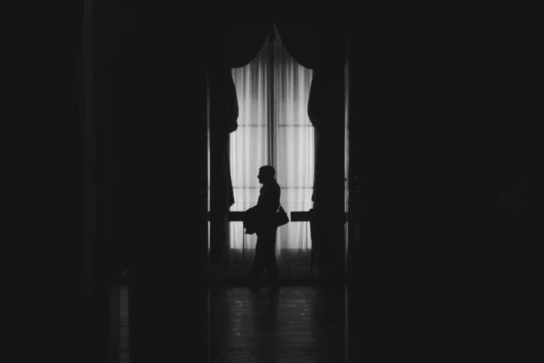 A figure silhouetted against a tall curtained window